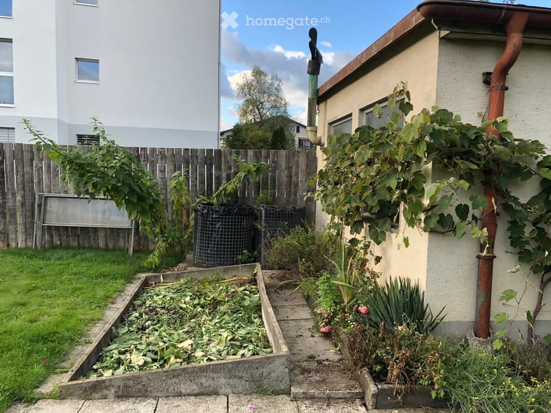 Garden area with a wooden fence, grassy lawn, raised garden bed, and various plants and shrubs. A water collection system is visible near the fence.