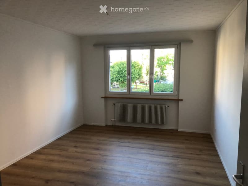 Empty room with wooden flooring, white walls, large window with a view of greenery, radiator, and a small wooden shelf under the window.