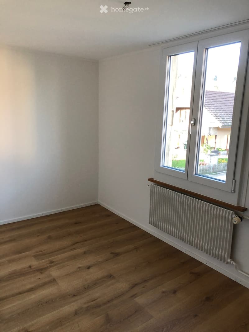 Empty room with wooden flooring, white walls, and a radiator under a large window offering a view of the outside.