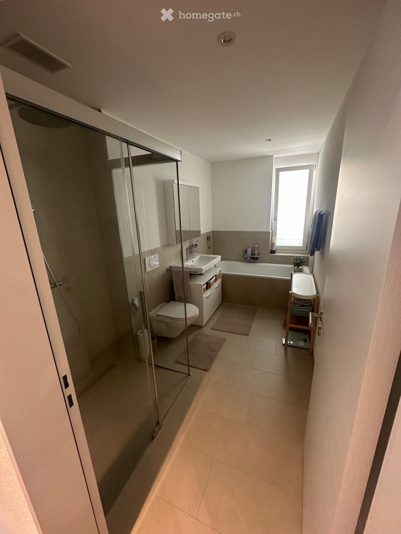 Modern bathroom with a glass-enclosed shower, toilet, sink, bathtub, and a small cabinet. Light-colored tiles and a window provide natural light.
