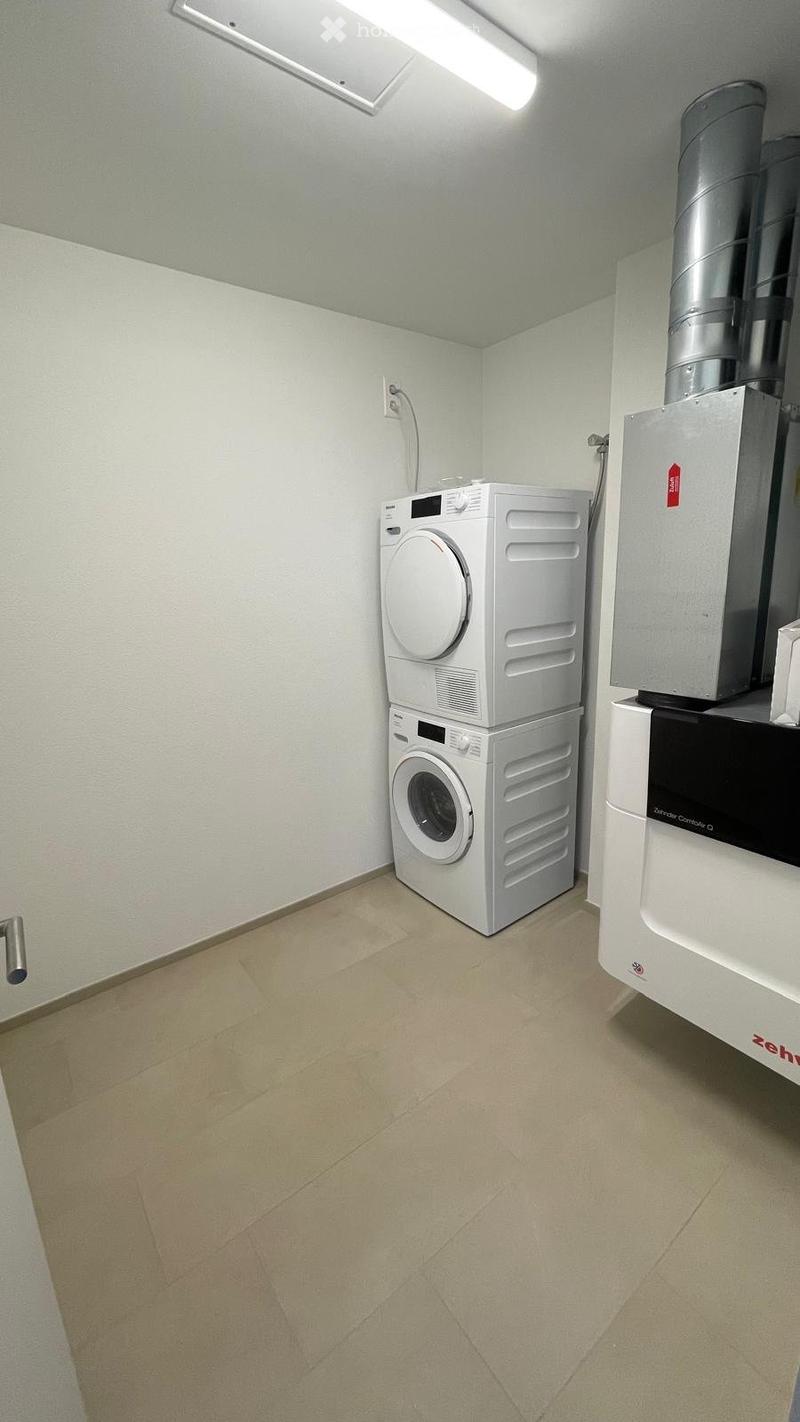 A small, clean utility room with a stacked washing machine and dryer, beige tiled floor, and white walls.