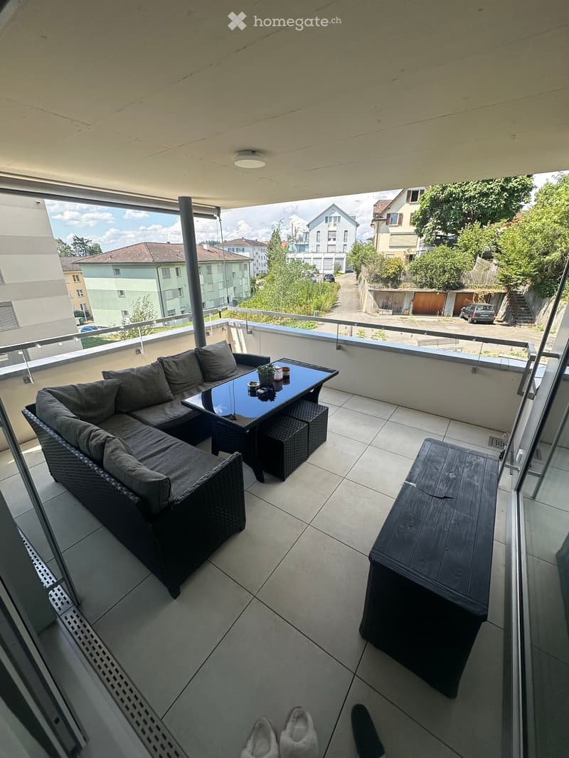 Balcony with a modern setup, including a dark grey sofa, a glass coffee table, two wicker stools, and a wooden bench, overlooking a residential area with houses and greenery.