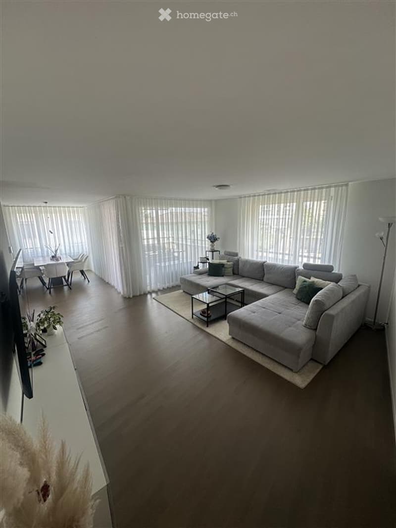 Modern living room with a large sectional sofa, coffee table, dining area with a table and chairs, large windows with sheer curtains, and a floor lamp.