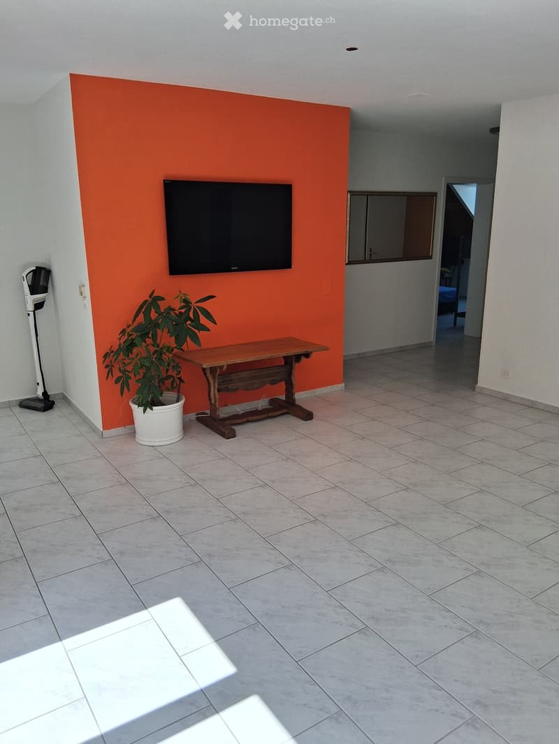 Living room with tiled floor, orange accent wall, mounted TV, small wooden table, potted plant, and a vacuum cleaner in the corner.