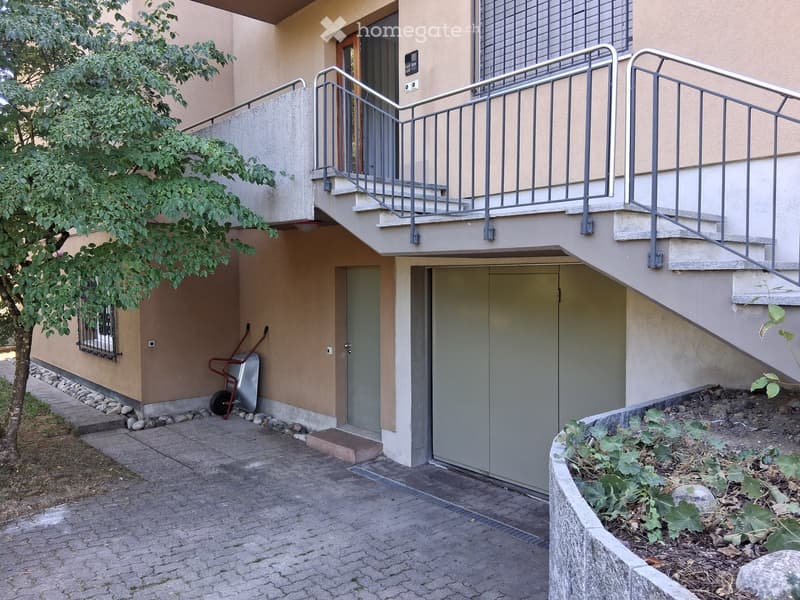 Two-story building with a raised ground floor, accessible via a ramp with metal railings. Features a garage entrance, small garden area, and a tree on the left side.