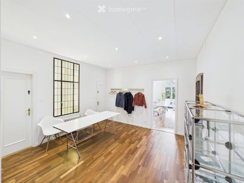 white walls, hardwood floors, two white chairs, long white table, hanging clothes on rack, glass case, radiator, window with stained glass