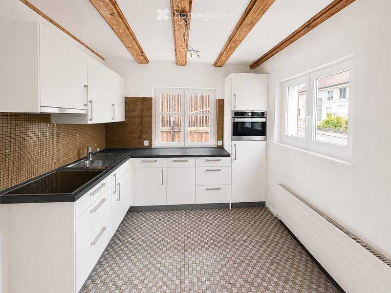Modern kitchen with white cabinets, black countertops, a sink, an oven, and a refrigerator. The space features exposed wooden beams on the ceiling and large windows offering a view of the outside.