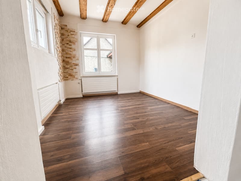 Empty room with wooden flooring, white walls, and exposed wooden beams on the ceiling. Features a large window with a white radiator below and a decorative wooden panel on one wall.