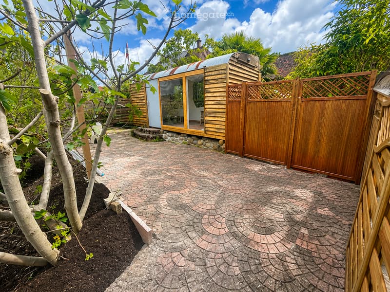 Garden area with a paved brick pathway leading to a small wooden cabin with a corrugated metal roof and glass walls, surrounded by trees and greenery.