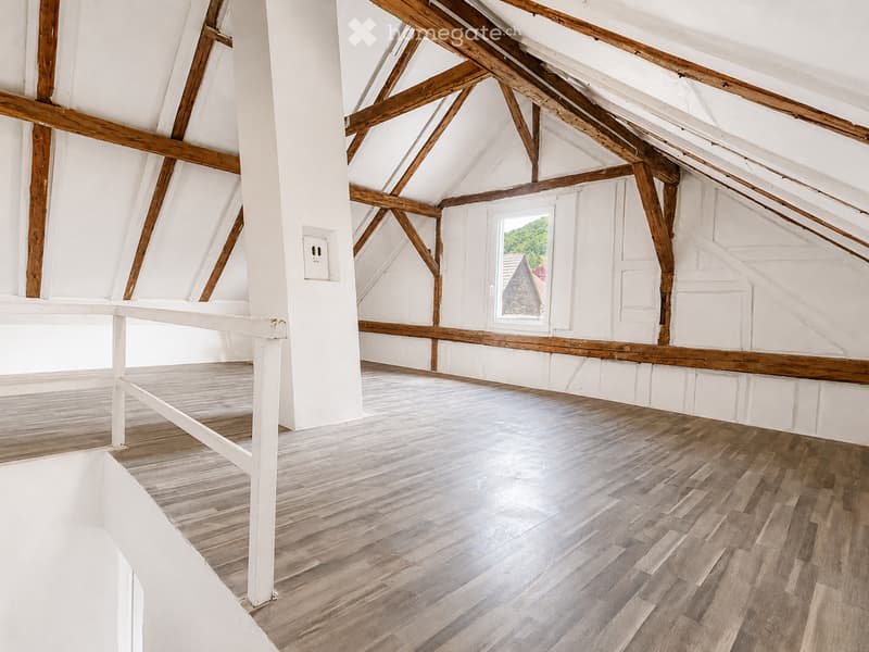 An empty attic space with exposed wooden beams, white walls, and wooden flooring. A single window provides natural light, and a white railing runs along the edge of the lofted area.
