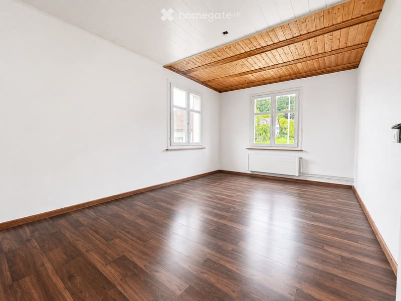 Empty room with wooden flooring, white walls, and large windows with white frames. The ceiling features exposed wooden beams and a white panel section.