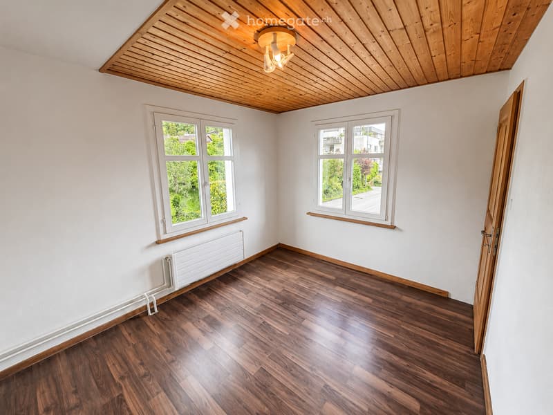 Empty room with wooden flooring, white walls, and large windows offering a view of greenery outside. The room features a wooden ceiling with built-in lighting and a radiator under one window.
