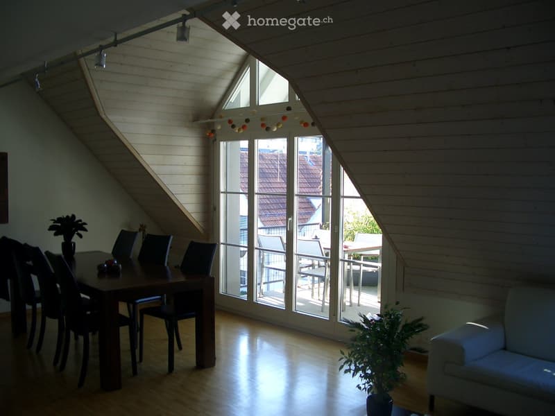 Bright, wooden-floored room with a slanted ceiling, large windows, and a sliding glass door leading to a balcony. Features a dining table with chairs, a sofa, and potted plants.
