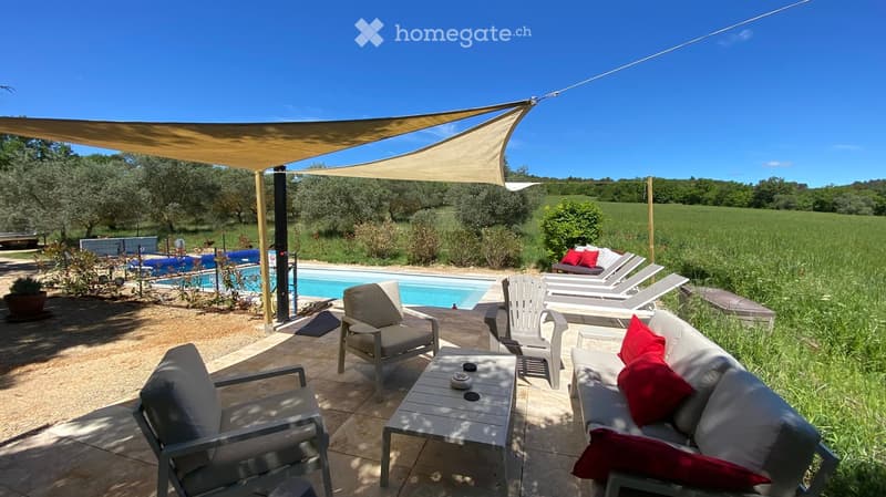 Outdoor patio area with lounge chairs, sofa, coffee table, and a swimming pool, surrounded by greenery and a large canopy.