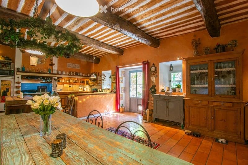A rustic, open-plan living and dining area with exposed wooden beams, terracotta tiles, and warm orange walls. The space includes a wooden dining table with chairs, a vase of white flowers, candle holders, a kitchen island with appliances, and wooden cabi