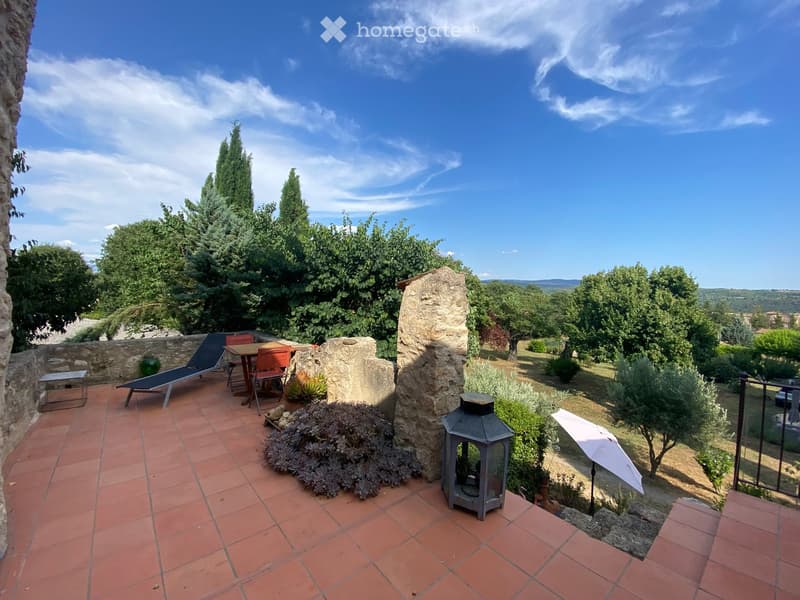 Terrace with red-tiled flooring, lounge chair, table, chairs, lantern, and umbrella, overlooking a lush garden and countryside.