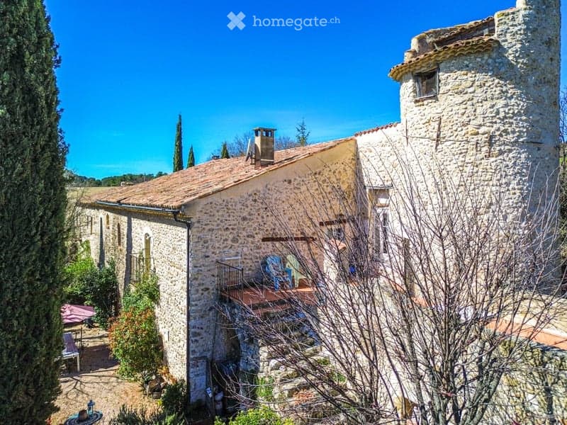 Stone house with terracotta roof, balcony, and surrounding garden area with outdoor furniture.
