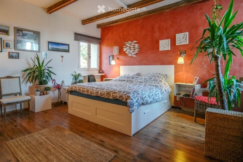 Cozy bedroom with wooden floors, a large bed with patterned bedding, two chairs, a small table, wall-mounted lamps, and several potted plants. The walls are painted in warm tones with decorative artwork.