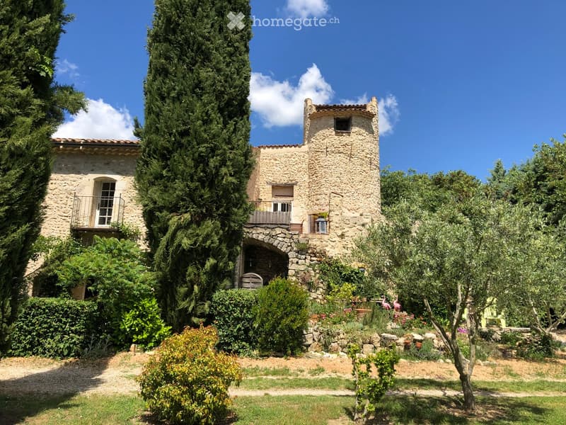 A rustic stone house with a tower, surrounded by lush greenery and trees, featuring a balcony and arched entrance.