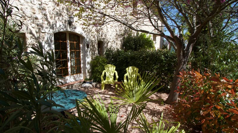 A serene garden area with a stone building in the background, featuring two green chairs, a blue table, and lush greenery including palm-like plants and flowering shrubs.