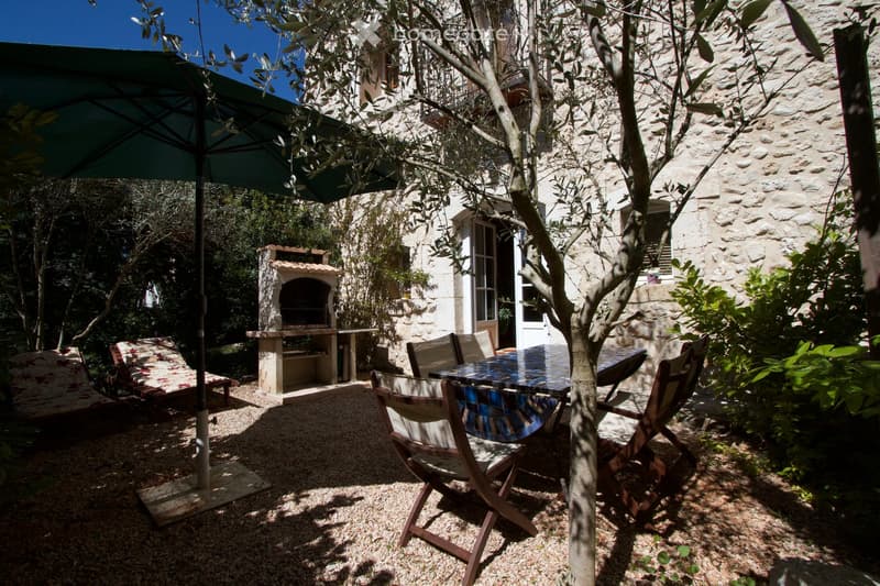 Outdoor patio area with a stone house in the background, featuring a table, chairs, a sun umbrella, a barbecue, and lounge chairs.