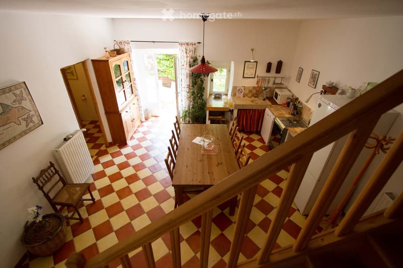A cozy, well-lit dining and kitchen area with checkered flooring, a wooden dining table with chairs, a kitchen counter with appliances, and a staircase leading upwards.