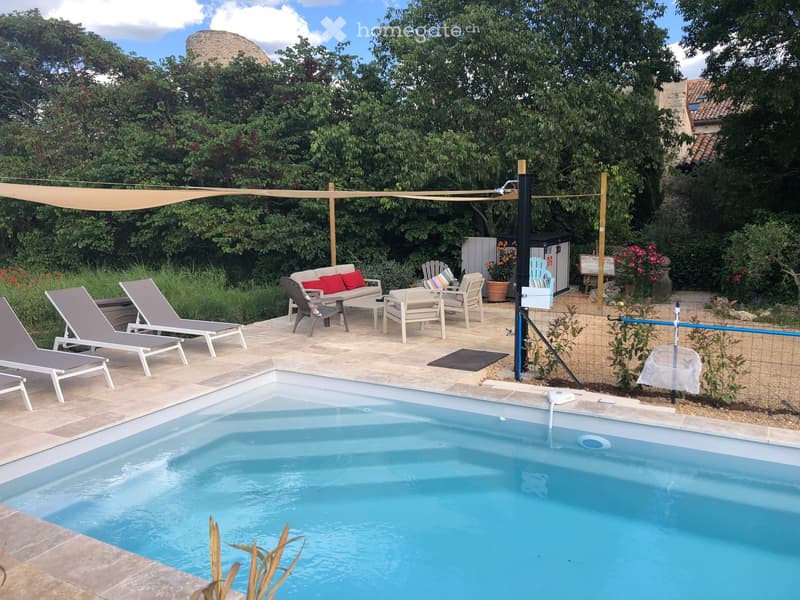 Outdoor patio area with a swimming pool, lounge chairs, a sofa, and a table surrounded by greenery and a pergola.