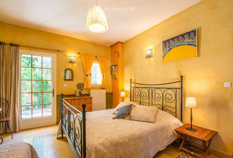 Bright bedroom with yellow walls, a large window with curtains, a wooden nightstand with a lamp, a metal-framed bed with pillows and a blanket, a small table with a vase, and a mirror on the wall.