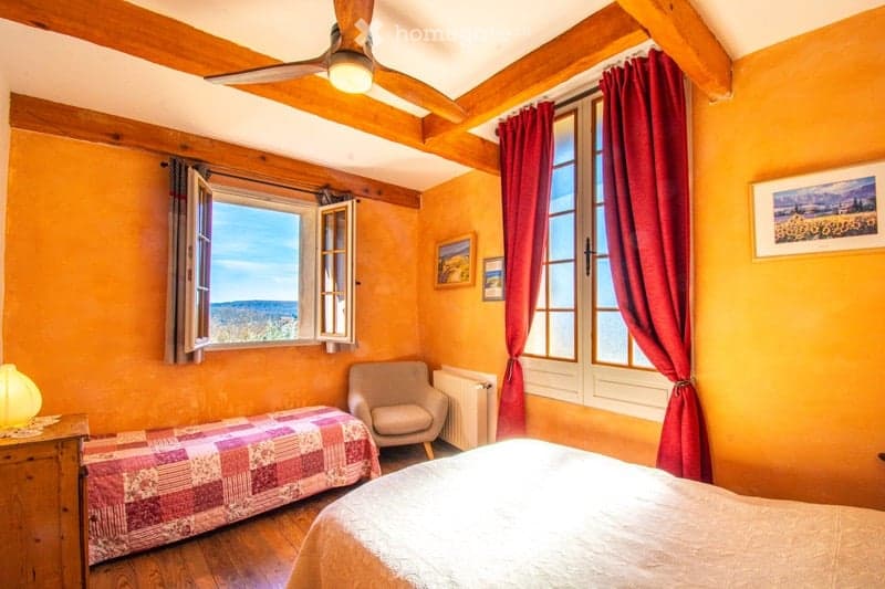 Cozy bedroom with exposed wooden beams, warm orange walls, large windows with red curtains, a bed with a patterned quilt, a wooden nightstand with a lamp, a single armchair, and a radiator.