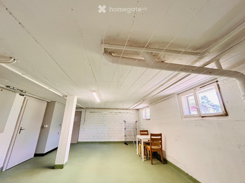 A small, unfurnished room with exposed brick walls, a table, and two chairs. The room has a green floor and white walls with visible pipes and wiring on the ceiling.