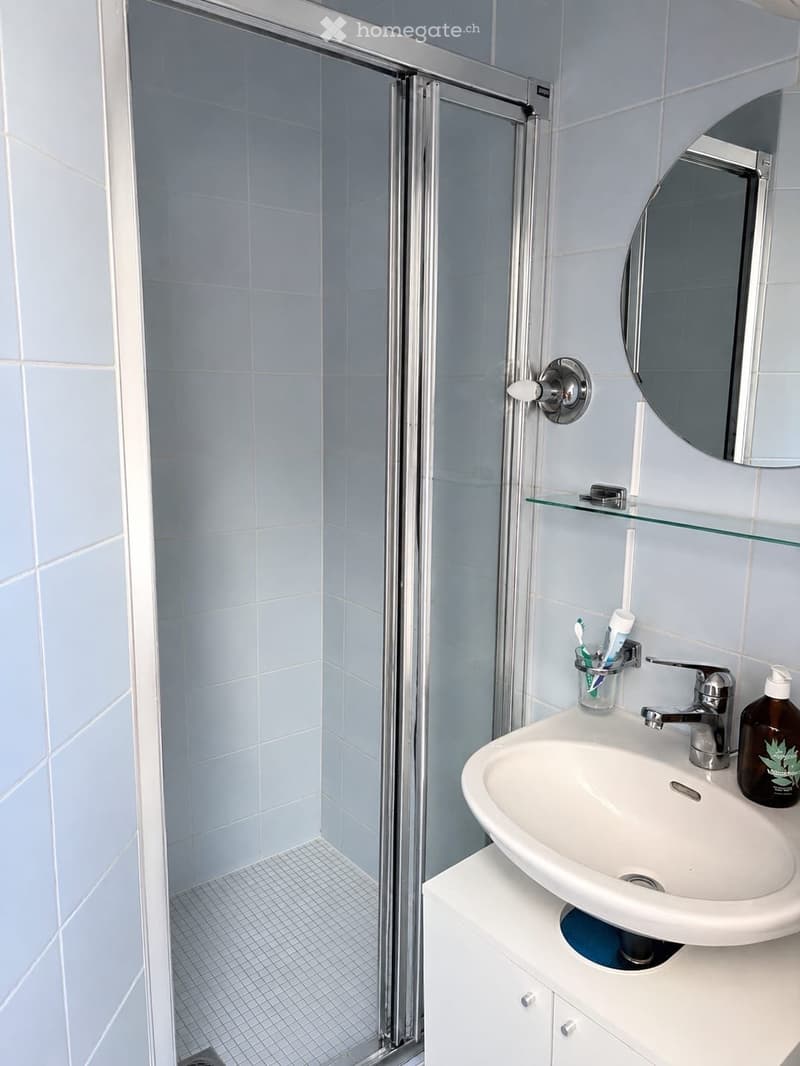 Modern bathroom with light blue tiles, a white sink with a chrome faucet, a mirrored cabinet, and a glass shelf above the sink. A toothbrush holder and a bottle are visible on the sink counter.