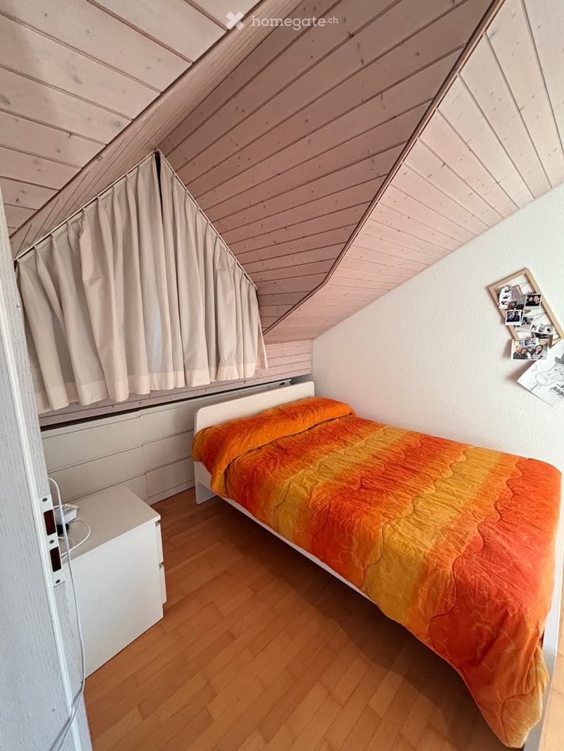 Cozy attic bedroom with wooden slanted ceilings, a single bed with an orange and yellow quilt, a nightstand, and a window with curtains.