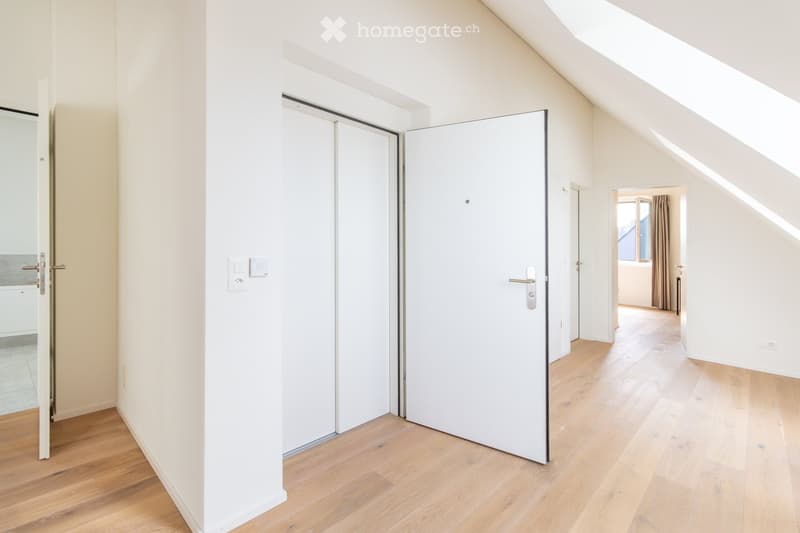 Empty hallway with light wooden flooring, white walls, and white sliding doors. Natural light streams in from a window at the end of the hallway.