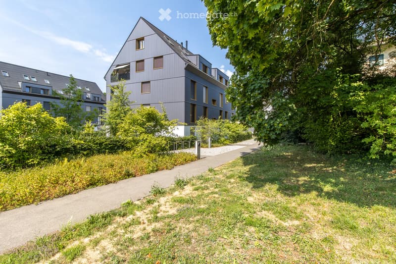 Modern residential building with grey facade and multiple windows, surrounded by greenery and a paved pathway.