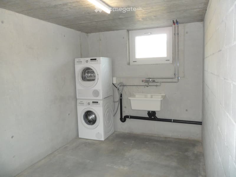 Concrete-walled room with a stacked washing machine and dishwasher unit, a small sink, and a window for ventilation.