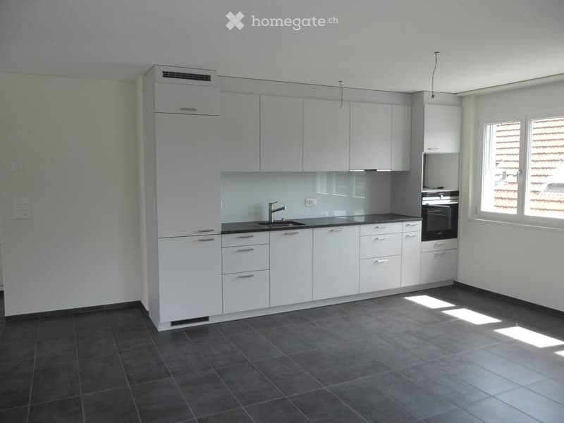 Modern kitchen with white cabinets, dark countertops, and a stainless steel sink. The space features built-in storage, a built-in oven, and large windows allowing natural light to enter.