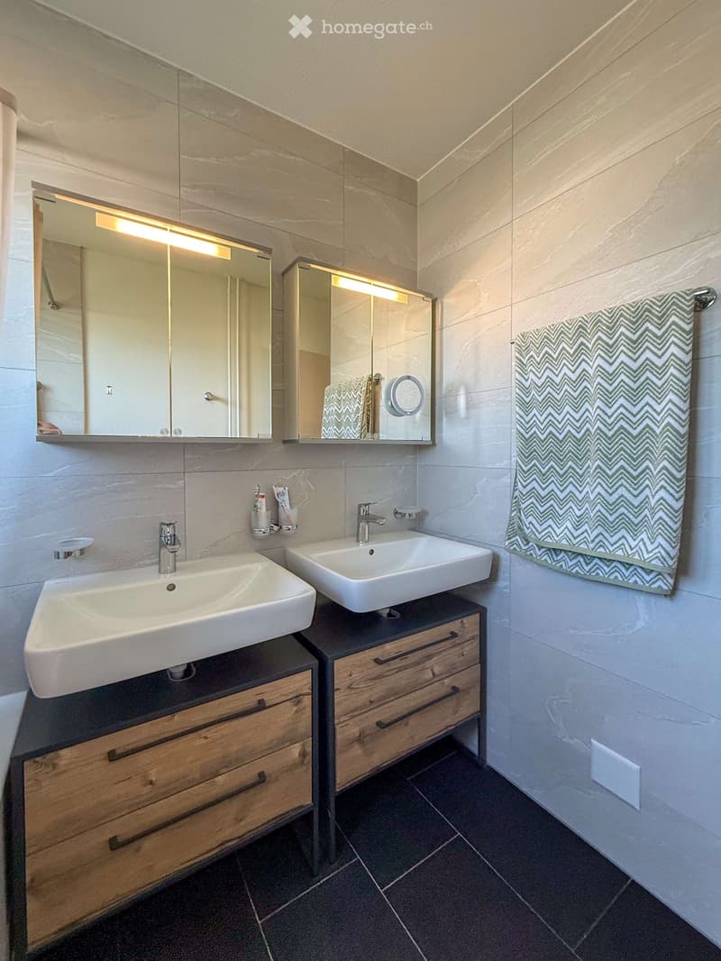 Modern bathroom with two white sinks, wooden vanity cabinets, mirrored cabinets with built-in lighting, and a decorative towel on the wall.