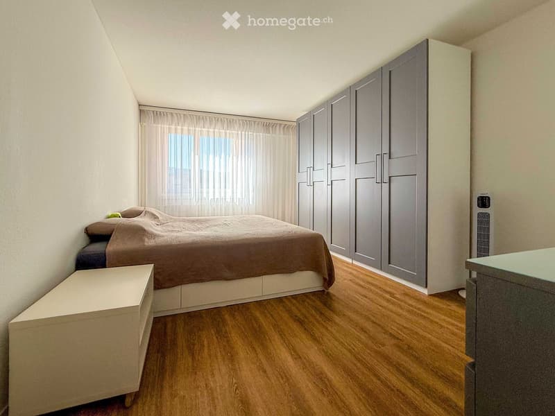 A modern bedroom with a large bed, a wardrobe, and a nightstand. The room features wooden flooring and large windows with curtains.