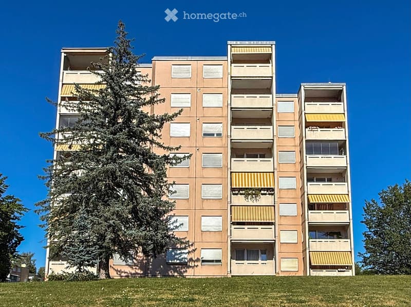 Multi-story residential building with balconies and yellow awnings, surrounded by trees and grass.