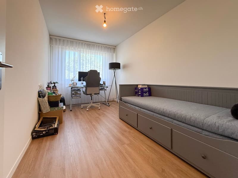 A modern bedroom with a bed, desk, chair, and lamp. The room features wooden flooring, large windows with curtains, and a variety of storage items.