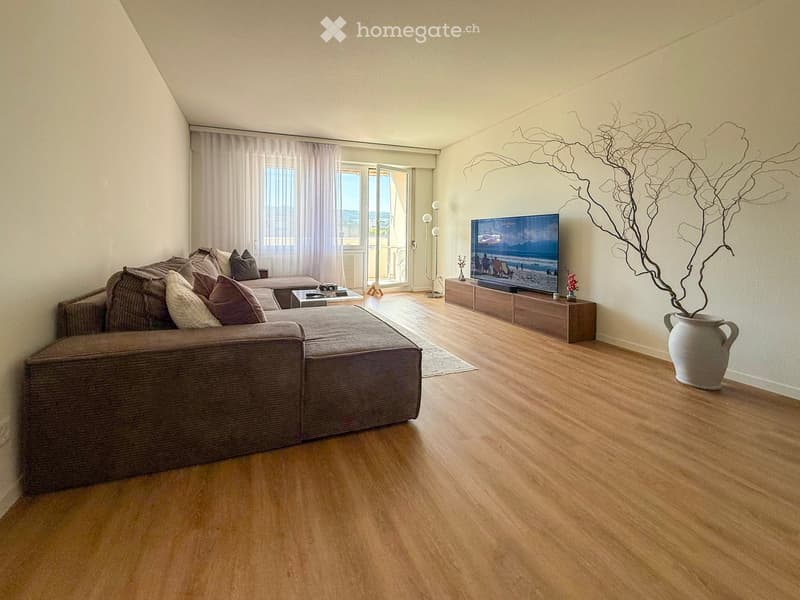 Modern living room with a sectional sofa, coffee table, flat-screen TV on a wooden stand, floor lamp, and decorative vase with dried branches.