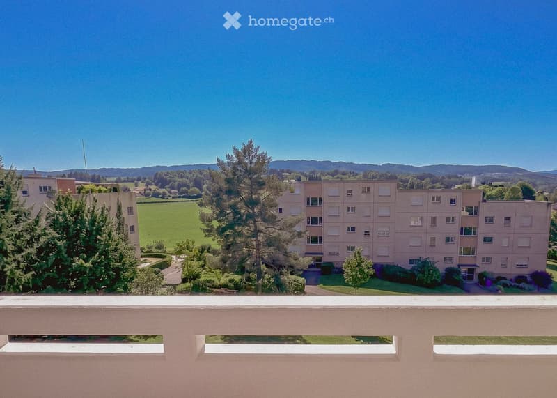View from a balcony showing a multi-story residential building surrounded by greenery and a scenic countryside with distant hills.