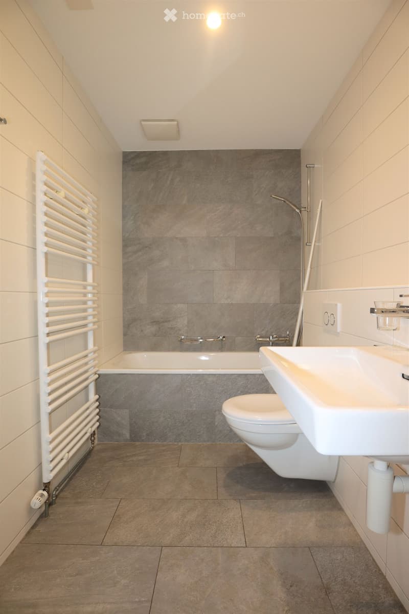 Modern bathroom with a bathtub, toilet, sink, and a white radiator. The walls are tiled with a dark grey accent wall behind the bathtub.