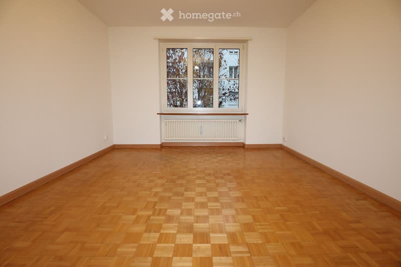 Empty room with light-colored walls, wooden parquet flooring, and a large window with white frames and a radiator below it.