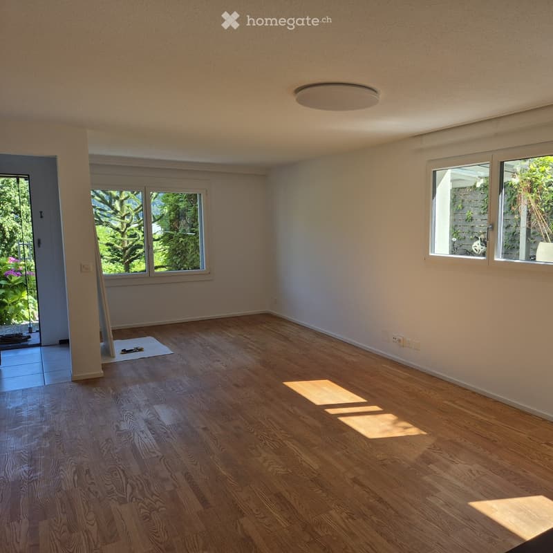Empty room with wooden flooring, large windows, and a sliding glass door leading to a garden area.