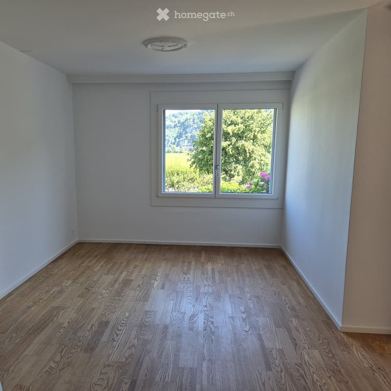 Empty room with wooden flooring, white walls, and a large window with a view of greenery and flowers outside.