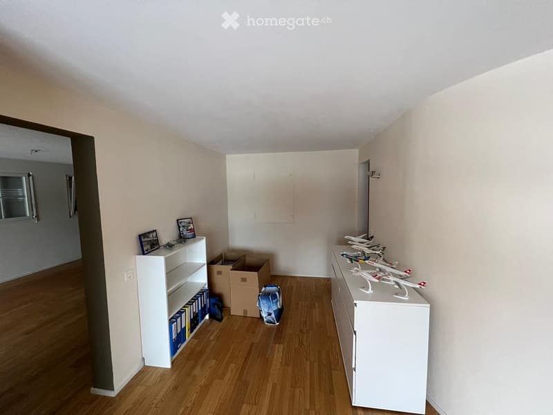 A small, unfurnished room with wooden flooring, white walls, and a shelf holding picture frames and books. A white dresser with toy airplanes is visible, along with cardboard boxes and a backpack on the floor.