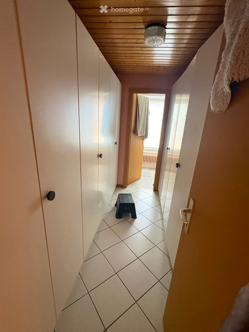 A narrow hallway with tiled flooring, white walls, and a wooden ceiling. A small black stool is placed in the center, and a towel hangs on the right wall. A door at the end leads to another room.