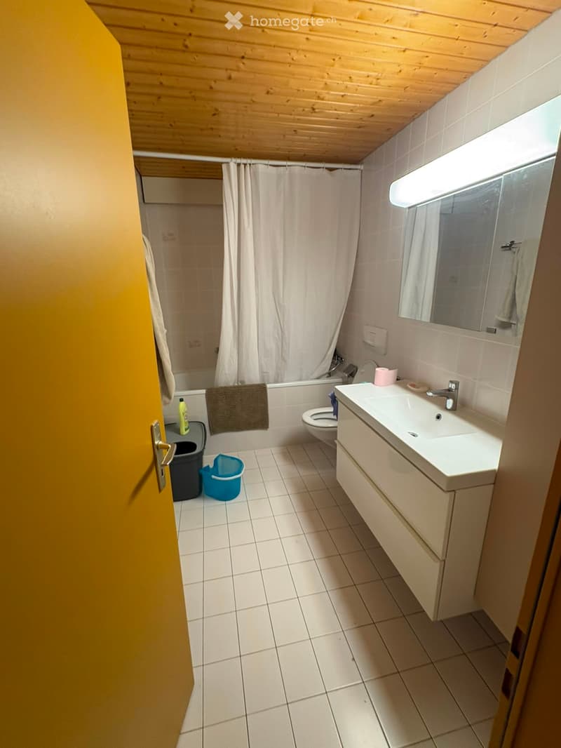 A small bathroom with white tiled floors and walls, featuring a sink, toilet, bathtub with a curtain, and a mirror above the sink. A towel hangs on the side of the bathtub, and a small trash bin is near the door.