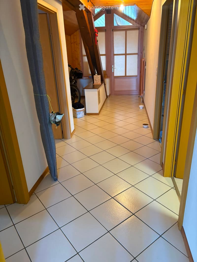 A narrow hallway with tiled flooring, wooden doors, and a small bench with a bicycle and items placed on it. The hallway leads to a door with glass panels at the end.